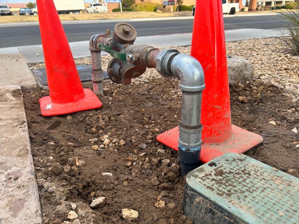 Water valve installation surrounded by orange safety cones on dirt surface near a road.