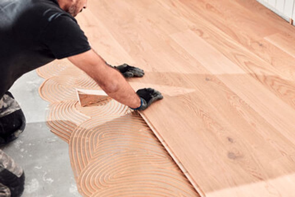 Man installing wooden flooring with adhesive, wearing gloves on a concrete surface.
